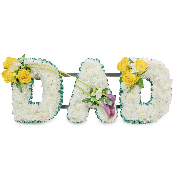 A deeply respectful floral tribute arranged to spell DAD in three large, freestanding capital letters, each letter composed of a dense bed of immaculate white spray chrysanthemums that lend a serene, cloud-like texture. Each letter's outline is crisply defined by a ruffled band of deep emerald satin ribbon, the dark green edge acting like a frame that enhances the luminous white blooms. Slim dark green metal bars run horizontally behind the letters and are visible through gaps in the flowers, indicating a sturdy connecting frame that keeps the trio aligned and ready for display. The leftmost D is accented on its upper left curve with a vibrant cluster of bright yellow roses-likely three or four open heads-nestled among tiny white filler blooms such as freesias or gypsophila and fresh green leaves; a pair of slender, bare green stems extend diagonally from that cluster toward the centre, giving the layout a gentle sense of motion. The central A is graced at its lower diagonal by two rich, deep-purple calla lilies, their sculpted cups offering an elegant, contemplative focal point surrounded by delicate white accents. The right-hand D repeats the cheerful yellow-rose motif on its upper right curve, mirroring the left for emotional balance. Set against a plain white backdrop under even lighting, the overall impression is one of quiet dignity and tactile softness - a fitting, handcrafted memorial piece to honour a father, assembled with the same care local families in New Barnet, from Monken Hadley Common to Oakleigh Road, have come to expect.