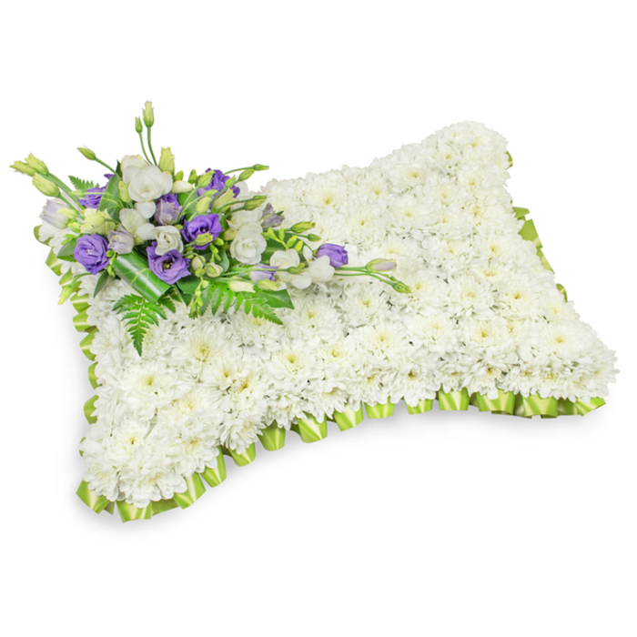 An elegantly composed rectangular pillow of remembrance, shown against a pristine white background with a soft shadow that suggests its slight puff and the careful handcrafting beneath. The surface is densely covered with hundreds of small white chrysanthemum heads, their tight, rounded petals forming a tranquil, uniform plane that reads as softness and serene repose. The entire perimeter is trimmed with a light lime-green satin ribbon, pleated into a gentle scalloped edge that defines the pillow's shape and brings a fresh, hopeful accent to the otherwise monochrome field. Resting on the pillow's upper-left corner is a tasteful accent spray: layers of white lisianthus and deeper purple lisianthus blooms-some buds still closed-nestle among feathery fern sprigs and broader dark-green leaves; a darker green wrap binds the cluster neatly at its base. This combination of crisp white and dignified purple offers a visual language of respect and remembrance, while the green foliage introduces texture and life. The studio lighting is soft and diffused, revealing subtle petal textures, the sheen of the ribbon, and the shadow that grounds the piece; every detail has been composed to convey calm and comfort. Suitable as a graveside cushion or chapel tribute, this arrangement evokes the quiet of a morning walk through Oak Hill Park or the hush of a service near New Barnet Station, acting as a compassionate floral message for those gathered to remember a loved one.