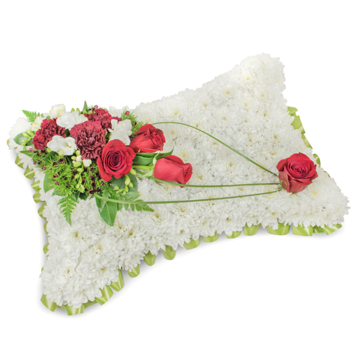 A carefully composed sympathy pillow occupies the frame: a low rectangular cushion entirely covered with small, pristine white chrysanthemum blooms whose tight heads create a sumptuous, fleece-like texture that suggests peace and repose. Around the slightly raised edge, a subtle strip of light green satin ribbon or finely gathered foliage peeps through, defining the silhouette with a fresh, lime hint. On the top-left corner, an artful spray of deep red flowers provides a strong, respectful accent-several rich red roses, some just unfurling and others fully open, sit alongside deep burgundy carnations with their ruffled, layered petals. Interspersed among these are clusters of small white flowers, likely freesias or spray chrysanthemums, which bring brightness and a delicate scent to the composition. A variety of green leaves and feathery fern sprigs nests behind and between the blooms, adding depth and grounding the arrangement. From that main cluster, graceful arcs of bright-green bear-grass or similar long foliage sweep across the white chrysanthemum surface toward the lower-right corner, culminating in a single, perfectly formed red rose that appears gently placed as the piece's visual punctuation. The lighting is soft and even, accentuating the contrast between the cool white cushion and the velvety crimson blooms while revealing subtle textures of petal and leaf. This dignified memorial arrangement-timeless in its white-and-red palette-would be fitting for funeral tributes around New Barnet, including services near the High Street, community chapels, and the surrounding residential streets where neighbours seek a composed expression of sympathy and enduring love.