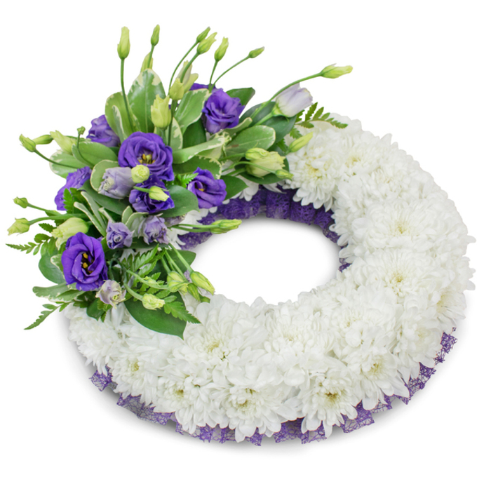 A round, thoughtfully crafted funeral wreath set against a starkly white background, defined by a thick, plush ring of immaculate white cushion chrysanthemums. The chrysanthemums are tightly massed to create a velvety, uniform surface whose layered petals suggest softness, purity and a sense of perpetual embrace. Positioned on the upper-left portion of the wreath is a vibrant focal cluster of purple Eustoma (lisianthus), their ruffled petals moving through a spectrum from deep, royal violet to softer lavender, intermingled with smaller buds that hint at unfolding life. Woven through the purple spray is an assortment of fresh green foliage - broad, variegated leaves and fine-textured fern-like sprigs - which adds verdant contrast and a subtle aromatic note. A delicate, crinkled purple mesh trims the inner and outer edges beneath the chrysanthemums, peeking out to provide a considered, tonal frame that unifies the composition. The arrangement is lit with soft, even lighting that casts a gentle shadow underneath, enhancing the wreath's three-dimensionality and handcrafted detail. As a local New Barnet florist I imagine this wreath being chosen for a wake or funeral service at a nearby chapel or community venue on Barnet High Street, offering a composed, respectful presence that conveys condolence and long-lasting remembrance without excess. The overall effect is one of calm restraint and floral craftsmanship, intended to comfort mourners and honour a cherished life.