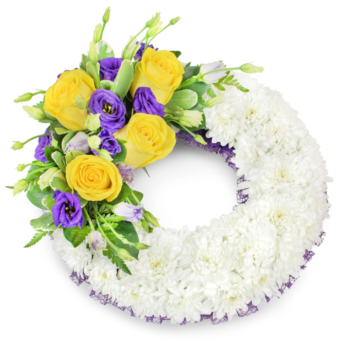 An expertly assembled circular sympathy wreath, shown on a clean white background, whose quiet palette and careful textures suit funeral use around New Barnet and the Oak Hill area. Its broad foundation consists of densely set white spray chrysanthemums, clustered so closely that the surface becomes a tactile, cloud-like continuum of delicate petals-soft to the eye and evoking reverent stillness. Anchoring the composition at the upper left is a colourful spray composed of several fully opened yellow roses with warm, buttery tones, contrasted by deep purple lisianthus whose ruffled, cup-shaped flowers bring a cool, dignified counterpoint. Pale lavender lisianthus buds and small green buds add soft transitional tones and a naturalistic feel, while glossy green leaves-some faintly variegated-and slender fern fronds weave between the blooms to create depth and structural support. A subtle dark violet mesh ribbon is visible beneath the chrysanthemum ring along both inner and outer edges, a delicate detail that repeats the purple accent and refines the overall silhouette. The wreath's open centre and balanced asymmetry allow for placement on an easel or over a memorial table, and its quiet scent of roses and fresh foliage would be faint yet comforting. As a local florist might note, this composition is appropriate for remembrance ceremonies across New Barnet High Street, Station Road, and nearby Brunswick Park, presented with considered craftsmanship and sympathetic intent.