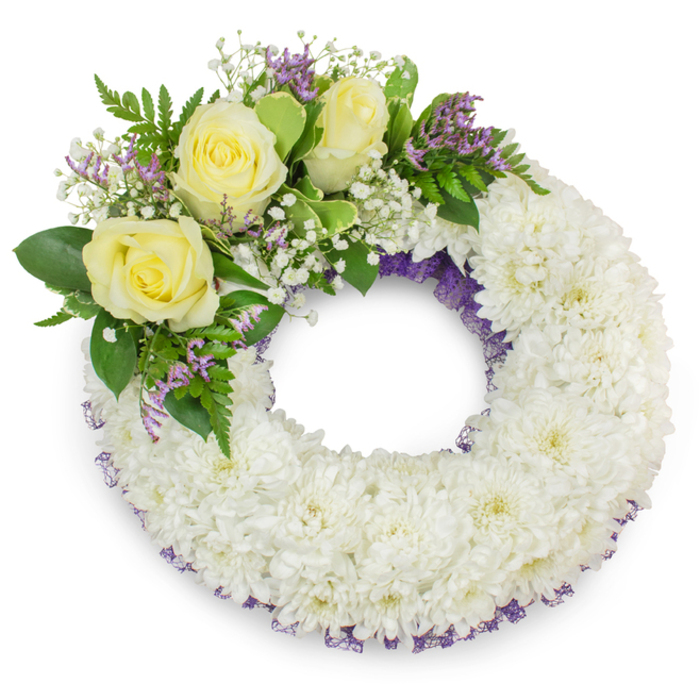 Presented against a stark white background, this circular sympathy wreath is a study in gentle contrasts and careful proportion. Hundreds of small white chrysanthemums are artfully arranged to create a sumptuous, even texture - their soft, layered petals forming a continuous band of white that reads as both pure and consoling. From the upper-left quadrant a composed cluster of accents introduces warm and grounding notes: three pale yellow roses bloom with layered, velvety petals that suggest closeness and lasting affection; clusters of delicate baby's breath settle among them like a soft halo; and tiny spikes of lavender-purple filler, likely statice or limonium, bring a modest pop of colour. Sprays of green foliage, ranging from broad, satiny leaves to slender, fern-like fronds, are interwoven to give the focal cluster movement and depth. A subtle ribbon or fine purple mesh is visible beneath the chrysanthemums along the inner and outer circumference, its muted colour lending a refined border without competing with the main blooms. The lighting is soft and neutral, emphasizing petal textures and casting a faint shadow that lifts the wreath from the background. This wreath reads as a respectful and elegant choice for a memorial, the kind of carefully tended tribute we prepare for families across New Barnet and surrounding areas such as Brunswick Park and Oakleigh - a tactile, visual gesture of compassion at a time of farewell.