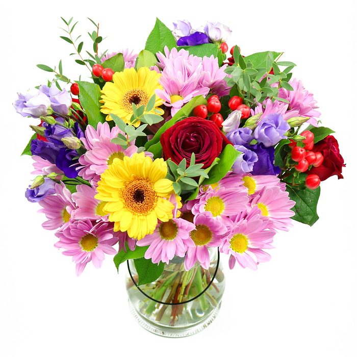 In the photograph, a lush mixed bouquet is presented in a clear glass vase, standing alone against a crisp white background that lets every detail of the flowers shine. The dominant impression is one of abundance and balance, with deep red roses anchoring the design at the centre, their plush, velvety petals adding a sense of intimacy and timeless romance. Around them, bold yellow gerberas, each with a pronounced dark brown-orange eye, radiate outwards like spokes of sunshine, giving the bouquet a cheerful, welcoming glow. Soft pink chrysanthemums cluster generously between the larger flowers, their many fine petals overlapping to create a pillowy texture that softens the vivid tones. Interwoven among the daisies and roses are slender stems of lisianthus in shades of rich purple and gentle lavender, their bell-like blossoms and slightly ruffled edges bringing a touch of modern elegance and visual depth. Here and there, small sprays of round red hypericum berries punctuate the arrangement, offering a playful, glossy contrast to the softness of the petals. Layers of green foliage-from broad, dark leaves to lighter, more delicate fronds-wrap and support the blooms, giving the whole piece a fresh, garden-inspired feel. Through the smooth glass sides of the vase, the neatly cut stems are visible, arranged with care in clear water. The bouquet exudes a bright, joyful energy, the kind you might send to a family home near Oakleigh Park or a new flat close to New Barnet high street to celebrate fresh starts, birthdays, or simple acts of kindness. It feels both thoughtfully composed and wonderfully spontaneous, like a little burst of New Barnet colour brought indoors.