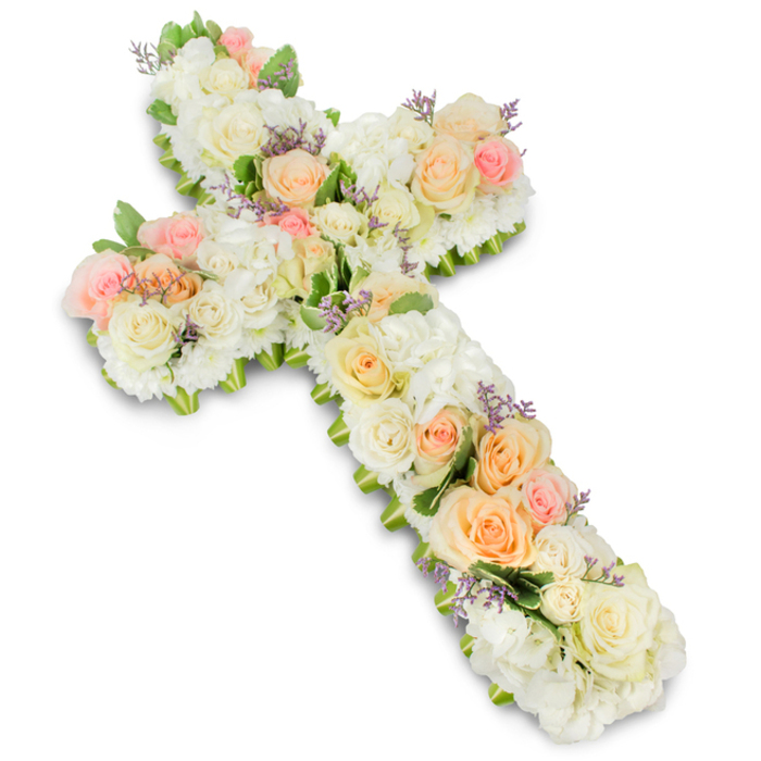Set against a crisp, brilliant white backdrop, a beautifully crafted floral cross dominates the image, symbolising both Christian faith and a gentle farewell. The cross is substantial in size and completely covered with fresh, meticulously arranged flowers, forming a luxuriant, almost quilted surface of petals. Soft white and ivory tones provide the main structure, with numerous roses fully opened to reveal their layered hearts, interspersed with equally generous clusters of fluffy white hydrangea or similar voluminous blooms. Together they create a plush, seamless base that traces the entire outline of the cross. Among this calm expanse of white, delicate pastel roses in tender shades of peach and light pink are artfully placed, creating small islands of colour that run along the length and arms of the design, giving rhythm and warmth to the overall composition. From certain angles, hints of pale lavender or light purple filler flowers emerge, adding a subtle, refined accent that fits harmoniously within the pastel palette. Around the outer edges, glimpses of fresh green foliage peek out, lightly framing the cross and adding definition, similar to how mature trees quietly frame the streets and gardens around New Barnet High Street. The blooms are arranged closely together, with no gaps, giving the impression of abundance and careful craftsmanship from a florist accustomed to preparing funeral tributes for local churches and crematoria. Soft studio lighting creates a gentle shadow beneath the cross, emphasising its depth and sculptural quality. The result is an image that conveys peace, remembrance, and enduring love-a dignified floral tribute that would feel at home at a memorial or funeral service in the New Barnet community.