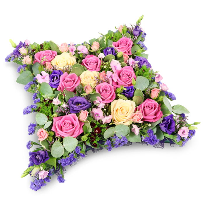 This image shows a beautifully crafted funeral cushion, designed as a soft rectangular pillow with rounded corners and displayed against a pure white background that throws every colour into relief. The floral surface is completely carpeted with blooms, giving the impression of a plush, petal-filled cushion resting in stillness. Dominating the arrangement are generous roses in three main shades: deep fuchsia pink, light blush pink, and creamy pale yellow, many of them fully open so their layered petals create a rich, velvety texture. Threaded throughout are elegant lisianthus in a deep royal purple, their ruffled, cup-shaped flowers adding a more dramatic note and enhancing the sense of depth from the centre outward. Scattered between the larger heads, clusters of tiny lavender and lilac filler flowers, likely statice or limonium, bring a fine, airy detail, while smaller spray roses in soft, romantic pink gently bridge the colour transitions. A border of mixed greenery wraps around and peeks through the blooms: rounded, silvery eucalyptus leaves introduce a cool, understated green, and slender, fern-like fronds offer darker accents, reminiscent of shaded corners in New Barnet's gardens. The cushion's compact, rounded form suggests comfort, repose, and a protective embrace, making it an especially meaningful piece for a funeral or memorial service. It feels fitting for a family gathering at a New Barnet chapel or crematorium, or as a carefully placed tribute at a local cemetery, where it would convey not only sympathy and respect but also a quiet promise of lasting remembrance and gentle, eternal rest.