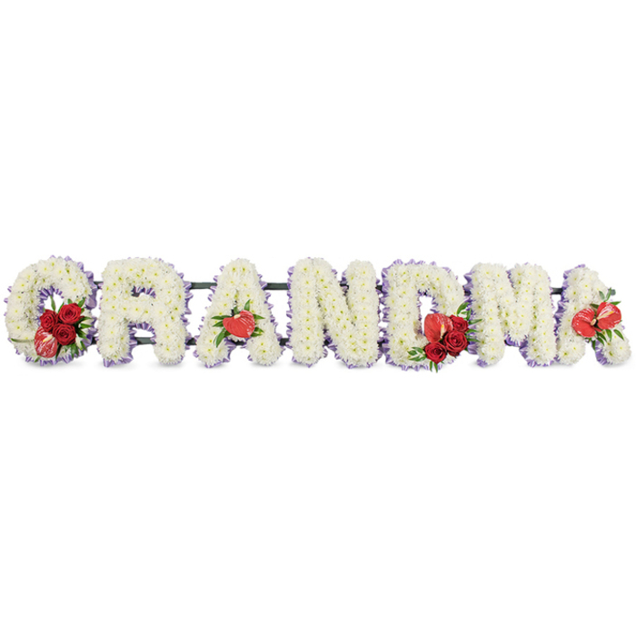 A carefully composed floral tribute spells out GRANDMA in bold, block capitals, arranged in a neat row and photographed against a simple white studio background that throws every detail into relief. The structure of each letter is completely filled with small white chrysanthemums or similar cushion blooms, packed tightly together so that no gaps show, creating a plush, almost velvety carpet of white. A soft trim of delicate purple ribbon or backing outlines each letter, gently tracing the shapes and lending a quiet hint of colour that makes the edges stand out. Throughout the tribute, rich red flowers are used sparingly but with clear intention. On the left, the letter G is adorned with a compact cluster of crimson roses entwined with a distinctive red-and-white variegated calla-like flower, positioned towards its base to draw the eye. The first A features a single, glossy red anthurium at its foot, the heart shape instinctively suggesting love. At the centre, the M carries another group of red roses, partnered again with a red anthurium that sits proudly among the white petals. To balance the design, the last A is completed with its own lone red anthurium. All the letters are mounted on a slim, dark green frame that runs behind them, providing support while remaining visually discreet. The tribute feels peaceful yet deeply affectionate, the kind of bespoke funeral arrangement a family in New Barnet might order for a grandmother's service at a local church off Station Road or for a quiet interment near Oakhill Park, conveying gratitude, memories, and a lasting sense of respect.