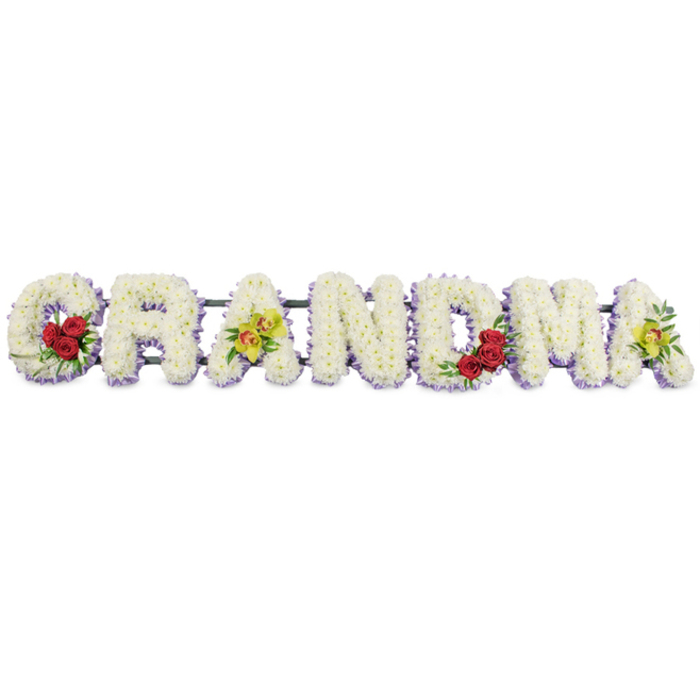 Set against a clean, white background, this photograph captures a full-length funeral flower tribute spelling out the word GRANDMA in large, neatly aligned capital letters. Every letter is meticulously shaped from masses of small, white chrysanthemums or similar tightly petalled blooms, packed so closely together that they form a continuous, velvety surface with a soft, cushion-like texture. Beneath the bed of white flowers, a fine underlayer of pale lavender or light purple blossoms peeks out, tracing the outline of each letter with a whisper of colour and lending depth and definition without disturbing the serene palette. On the left side, the G is accented at its lower curve with a compact group of three deep red roses nestled in dark green foliage, the roses' rich, velvety petals symbolising long-held love and respect. The first A is adorned across its midsection with two yellow-green cymbidium orchids, each bloom marked with a reddish-brown patterned throat, bringing an elegant, slightly exotic touch that feels personal and carefully chosen. The central letters R, N and D remain unembellished aside from the lavender edging, their simplicity reinforcing a sense of calm and continuity. Further along, the M carries another trio of red roses at its lower right, mirroring the sentiment of the opening letter, while the final A echoes the first with another pair of green orchids resting near its middle. The entire sequence of letters is fixed to a slim, dark metal framework that runs horizontally along the back, providing stability while remaining visually discreet. This GRANDMA tribute would sit beautifully beside a coffin or on a stand at a New Barnet funeral home, offering a tender, visually striking expression of remembrance from grandchildren and family members.