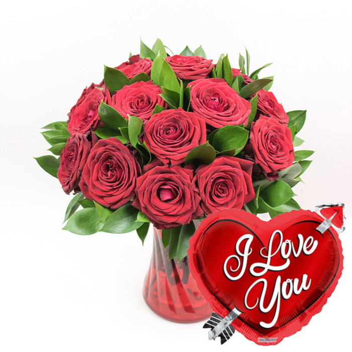 An opulent cluster of deep crimson red roses spills elegantly from a clear, fluted ruby-red glass vase, positioned against a pristine white backdrop that lets every detail stand out. The bouquet features roughly a dozen or more roses, their petals soft and velvety, arranged so closely that they form a rich, almost cloud-like dome of colour. Some blooms are wide open, revealing spiralled centres that draw the eye inward, while others remain partially closed, promising to unfurl over the coming days. Interspersed among the flowers are glossy dark green leaves, likely sturdy ruscus foliage, which weave through the arrangement and create a vibrant contrast, much like a patch of green in New Barnet's quiet residential streets. The vase beneath is a clear glass form tinted in a ruby hue, with subtle vertical ridges that catch the light, offering texture and a sense of height to the composition. Just in front and to the lower right of the bouquet, a shiny red heart-shaped foil balloon leans into the frame. Its metallic surface reflects highlights and soft reflections from the surrounding white space. Across the balloon's centre, in sweeping white cursive script, the words I Love You stretch boldly, easy to read yet romantic in tone. A silver Cupid-style arrow seems to pierce through the heart, entering at the lower left and exiting at the upper right, reinforcing the idea of being love-struck. The overall image feels like a carefully prepared romantic gift, the kind of arrangement that could arrive as a surprise same-day delivery for a partner in New Barnet or nearby Barnet town centre, perfect for anniversaries, proposals, or Valentine's Day celebrations.