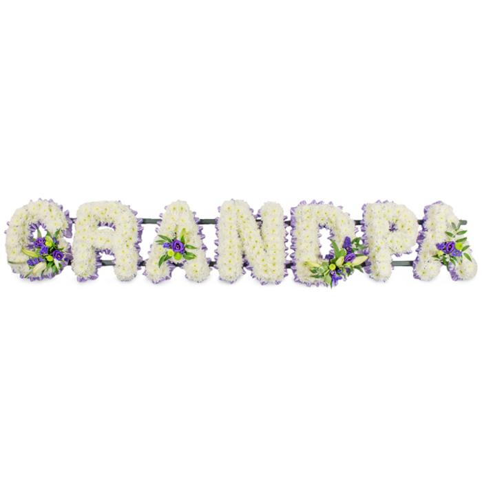 Spanning the width of the image, the word GRANDPA is beautifully rendered in large floral lettering, each capital letter formed entirely from fresh white blossoms, arranged horizontally against a spotless white backdrop that emphasises their clean lines. The small white chrysanthemums or carnations are meticulously layered, petal upon petal, so that the letters look almost sculpted from soft snow, with a plush, cushion-like texture. Every surface is covered, from the inner curves of the 'G' to the straight edges of the 'N', giving the tribute a polished, professional finish that reflects the care of an experienced funeral florist familiar with services in New Barnet churches and chapels. Interwoven through the white base are subtle bursts of colour: clusters of deep purple and lavender flowers, complemented by glossy green foliage. These accents nestle thoughtfully at key points-the base of the 'G', the centres of the 'A's, within the rounded parts of the 'D' and 'P', and enhancing the final letters-creating a gentle rhythm of colour that leads the gaze across the name. The contrast between the cool, tranquil whites and the richer purples suggests both sorrow and enduring love, echoing the mixed emotions of loss and gratitude at a memorial gathering. Beneath the flowers, a dark green metal or foam-backed support bar discreetly connects each letter, hidden under the blooms but essential for keeping the tribute straight and secure, whether displayed at New Barnet crematorium or carried from a hearse to the graveside. Overall, the arrangement conveys a strong sense of devotion to a much-loved grandfather, offering a personalised, visually striking way for a family to express Grandpa in flowers at a final farewell.