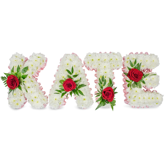 A refined, handcrafted lettering tribute reads KATE in substantial block forms, each letter composed entirely of compact white chrysanthemums whose small blooms interlock to create a plush, sculptural surface. Set against a plain white backdrop, the purity of the white chrysanthemums is emphasized, and the letters read like soft, tactile panels. Along the outer contours a pale pink satin ribbon has been applied as an elegant border, its subtle colour and sheen introducing a gentle contrast that frames the floral shapes and enhances their three-dimensional quality without overwhelming the composition.
Each letter is embellished with one vivid red rose, its deep crimson tones creating an intentional focal point amid the white. The roses are cradled in an arrangement of green foliage - a combination of broad, glossy leaves and fine, fern-like sprigs that add texture, shadow and natural movement. Attention to placement is precise: the rose on the K is set on the lower portion of its left vertical stroke; the A's rose crowns the top of its triangular peak; the T's bloom nestles at the base of the left side of its horizontal bar; and the E's rose is positioned on the middle horizontal arm. This personalised floral name tribute is typical of bespoke sympathy work - suitable for a funeral service or celebration of life - and reflects the kind of considered, detail-rich floristry often requested by families in New Barnet for services near Victoria Recreation Ground or the local church halls.