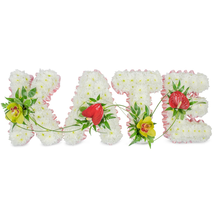 Set against a bright, plain white background, a personalised funeral flower tribute spells out the name KATE in large, clearly defined letters, as if laid out for a service in one of New Barnet's peaceful chapels. Each capital letter is constructed from tight clusters of white chrysanthemums, the small, multi-petalled blooms forming a dense, plush surface that appears as soft as a pillow. Their uniform white tone conveys simplicity and purity, while the slight variations in petal shape give the tribute a living, organic feel. Around the edge of every letter runs a neat frame of light pink ribbon, gently ruffled, which lends a feminine charm and helps each character stand out crisply against the background. Within this snowy base, brighter accent flowers and foliage provide contrast. A yellow-green cymbidium orchid with a speckled reddish-brown centre nestles within the K, mirrored by another on the T, both surrounded by glossy dark green leaves that add depth and structure. The A and E feature striking, heart-shaped red anthuriums with a satin-like sheen, their vivid colour symbolising love and remembrance. Fine green tendrils and fern-like sprigs weave through and around the letters, visually connecting them so that the name feels like one continuous, flowing tribute rather than four separate pieces. The careful balance of colour-the whites, soft pinks, fresh greens, and rich reds-creates a calm, dignified arrangement that would sit beautifully beside a coffin or memorial display at a New Barnet funeral home. It is a thoughtful, custom-made floral expression of farewell to Kate, combining skilled craftsmanship with a tender sense of personal remembrance.