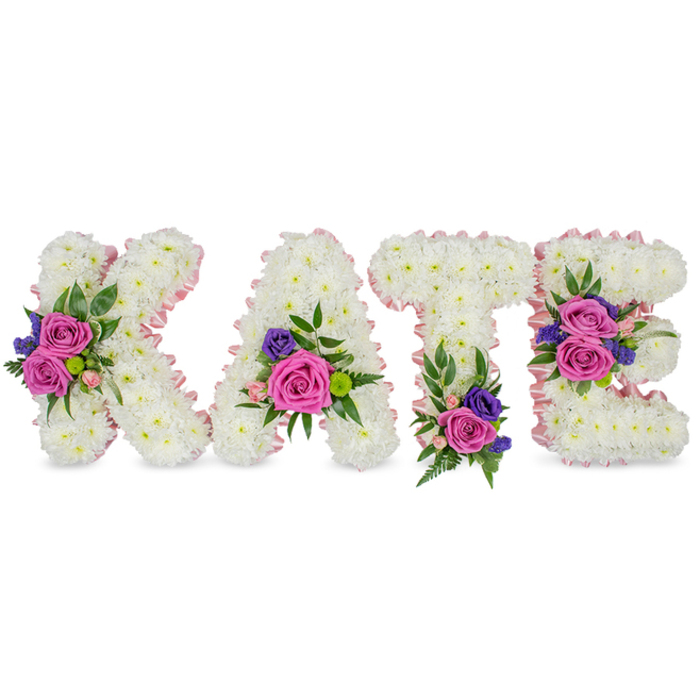 A solemn yet lovingly made floral name tribute spelling KATE, presented as four individual, free-standing letters fashioned entirely from compact clusters of immaculate white chrysanthemums. The chrysanthemums are arranged so densely that each letter reads as a smooth, textured block of white, the tiny petals forming a tranquil, cloud-like face on K, A, T and E. Each letter is edged with a pale pink satin ribbon that both outlines and softens the geometry of the characters, lending a gentle, compassionate note to the overall palette. On top of this serene foundation sit clusters of rich fuchsia-pink roses and smaller pastel-pink spray roses, their rounded petals adding warmth and a quiet perfume; small deep purple or navy-hued accents-probably statice or petite lisianthus-are used sparingly to introduce a measured contrast. Lacy fern fronds and broad eucalyptus leaves are threaded through the arrangements, contributing layered textures and a sense of natural depth. The decorative groupings are placed with thoughtful symmetry: the K's diagonal arm hosts a prominent cluster, the A is balanced with flowers near its lower right curve, the T carries a substantial base spray, and the E is graced by two floral clusters on its middle and lower bars. Set against a bright white backdrop with clean, flattering light, this composition reads as a dignified memorial piece perfect for a funeral, celebration of life, or grave-side service. As a florist who knows New Barnet's quieter traditions-from services at the parish church to small family wakes near Victoria Recreation Ground-this arrangement offers a personal and respectful way to honour a loved one named Kate.