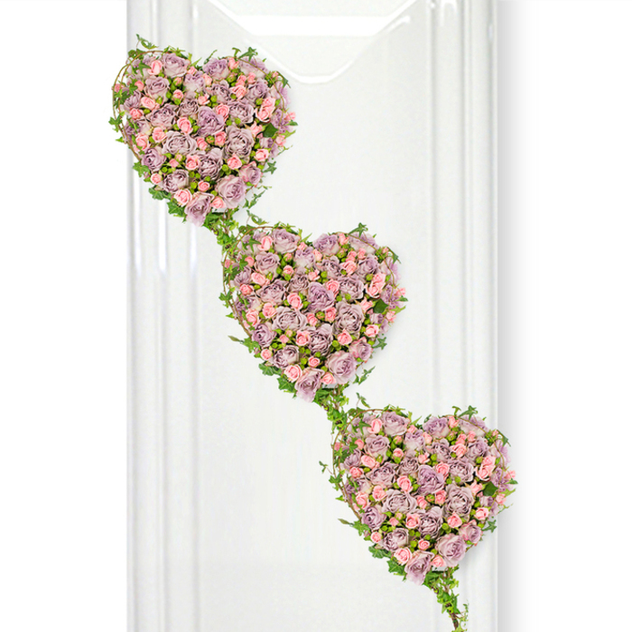 This image shows a beautifully composed triple-heart funeral tribute, crafted from delicate roses that seem to glow softly against a pure white background, like a moment of stillness amid the everyday bustle of New Barnet. Three hearts, each distinct yet seamlessly connected, cascade gently from top left to bottom right, giving the arrangement a flowing, story-like quality. The hearts are lavishly filled with small, tightly layered roses in shades of dusty lavender and soft mauve, accented with petite pastel pink roses nestled between them. The colours blend harmoniously, offering a calm, romantic palette that feels both modern and timeless. Every heart is traced and connected by strands of vibrant green ivy, the slender stems looping around the edges and then sweeping down to link one heart to the next. The ivy's glossy leaves provide a fresh, garden-like contrast to the velvety petals, suggesting resilience, growth, and continuity. Subtle translucent vertical lines on the left hint at a reflective surface or glass partition, adding a sense of depth without intruding on the gentle beauty of the flowers. The light is bright yet soft, highlighting the curves of each petal and the natural variations in tone. This tribute would sit gracefully at the front of a chapel or beside a framed photograph at a memorial in New Barnet, perhaps after a quiet family gathering near Oakhill Park. It evokes a message of three hearts beating as one-perfect for honouring parents, partners, or generations of loved ones whose memories remain closely entwined.