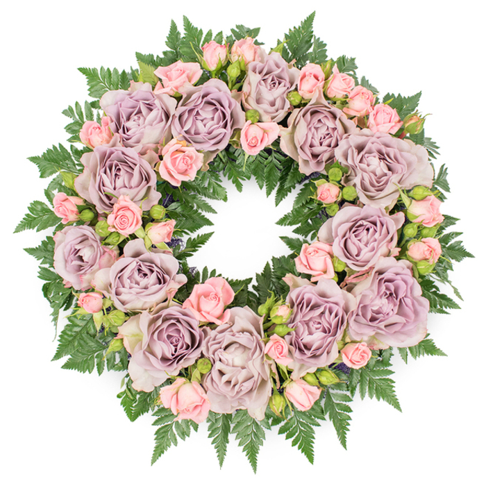 A meticulously arranged circular funeral wreath is shown laid flat on a crisp white surface, its form perfectly round and full, as though freshly crafted in a New Barnet flower studio just off East Barnet Road. The wreath is lavishly adorned with large roses in a sophisticated spectrum of dusty lavender, antique mauve, and soft, greyish-purple, each bloom fully open and displaying layers of velvety petals spiralling towards a gentle, shadowed centre. These stately roses are interlaced with clusters of smaller blush pink spray roses and petite rosebuds, some just beginning to unfurl. Their lighter tone and varied stages of opening give the design a sense of tenderness and gentle movement, like fond memories surfacing one by one. Beneath and around the blooms lies a dense collar of rich, dark green fern fronds, with fine, serrated leaves that extend beyond the flowers to trace the outline of the wreath. This deep greenery, possibly leatherleaf fern, provides both structure and contrast, making the soft pinks and muted purples appear even more serene. Between the main flowers, small hints of darker foliage and tiny filler buds create depth, suggesting the careful hand of an experienced florist. The lighting is bright but soft, casting no harsh shadows, allowing every texture-from the satin-soft rose petals to the slightly matte greenery-to be clearly seen. The colour harmony and balanced composition evoke a mood of peace, remembrance, and quiet dignity, making this wreath a fitting tribute for a funeral, cremation, or memorial service in the New Barnet area, whether delivered to a chapel near the station or a gathering in the calm surroundings of a local cemetery.