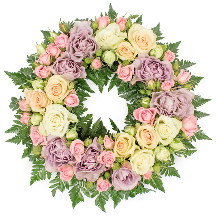 This image shows a graceful circular funeral wreath, photographed head-on against a spotless white background so that every detail of the arrangement stands out clearly. The wreath is abundant and substantial, completely cloaked in roses and foliage, with no bare spaces, as if it has been lovingly built petal by petal in a New Barnet flower shop. At first glance, the eye is drawn to the larger roses in calm, muted shades: creamy white blooms with softly cupped petals, pale peach roses glowing gently at the edges, and distinctive dusty lavender or mauve roses that bring a subtle, almost antique character to the composition. These big blossoms are carefully interwoven with clusters of smaller pink spray roses and pale green rosebuds, some tightly furled, others just starting to open, creating intricate detail and textural contrast reminiscent of the layered greenery in Monkfrith Open Space. The outer and inner edges of the ring are softened and framed by dark green fern fronds, their feathery leaflets emerging between the flowers in small arcs and points. This foliage adds depth, coolness, and a sense of natural woodland tranquillity, while its deep colour enhances the delicacy of the pastel tones. Soft, even lighting reveals the subtle variations in each petal-from ivory to cream, blush to peach, lavender to muted mauve-without casting harsh shadows, giving the wreath an almost luminous quality. The perfect, unbroken circle symbolises eternal love and remembrance, making the piece fitting for a chapel service, cremation, or graveside ceremony in or around New Barnet. Overall, the design exudes quiet dignity, gentle sorrow, and a comforting promise of lasting memory.