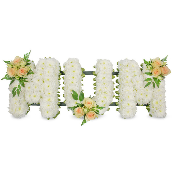 This photograph captures a lovingly made floral tribute spelling out MUM in large, upright capital letters, perfectly suited to rest along the side of a coffin or on a stand at a memorial service in New Barnet. Each letter is entirely filled with tightly clustered, pure white spray chrysanthemums, their small, daisy-like blooms overlapping to create a pure, even surface that feels soft and comforting. The chrysanthemums are set into a hidden frame, with a fine band of dark green ribbon or foliage glimpsed at the edges, crisply defining the contours of the M, U, and final M without distracting from the gentle whiteness of the flowers themselves. To break the white and bring a tender warmth, three small but expressive clusters of roses are carefully placed along the tribute. At the upper left shoulder of the first M, a mix of open roses and buds in soft peach and delicate blush pink spills over the chrysanthemums, surrounded by fronds of fern and slightly broader, deeper green leaves. The second rose cluster nestles at the lower centre of the U, drawing the eye to the heart of the word and adding a quiet, grounding note. The third sits on the upper right shoulder of the last M, completing a diagonal line of colour that feels both intentional and harmonious. The overall composition is simple yet rich with feeling, conveying love, gratitude, and remembrance. It is the kind of bespoke sympathy tribute a local family might order from a New Barnet florist who understands how important it is to say goodbye with beauty and care.