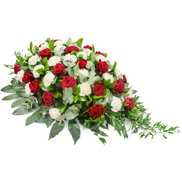 This image presents a graceful casket spray, photographed against a clean white background so that every detail of the design is clearly visible. The arrangement is horizontally oriented, with a lush, mounded form that is noticeably fuller on the left side and gradually narrows into a flowing point at the right, lending it an elegant, almost teardrop-like silhouette. Dominating the spray are deep red roses, each rosebud tightly formed with many velvety petals that suggest intensity of feeling and long-held affection. Between and around them, numerous white carnations open in soft, ruffled layers, their pristine colour providing a peaceful, contemplative contrast to the richness of the roses. Supporting the flowers is a generous layer of foliage: broad, dark green leaves create a strong base that holds the composition together, while scattered stems of silvery-green eucalyptus bring a cooler note and hint at their characteristic fresh, resinous scent. Toward the right-hand edge, fine, trailing greenery-resembling ruscus-extends out in small arcs, giving the arrangement a sense of natural movement and preventing the outline from feeling too rigid. The interplay of textures, from the velvet of the roses to the frill of the carnations and the smooth gloss of the leaves, adds depth and interest even in a still photograph. The traditional red and white colour scheme is perfect for a New Barnet family wishing to honour a loved one with something both classic and heartfelt, whether at a church service off the High Road or a quieter farewell nearby. The spray appears ready to be placed on a casket or memorial stand, offering a composed, dignified expression of sympathy and remembrance.