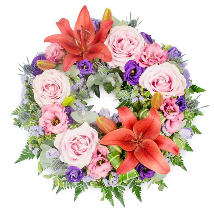 A beautifully composed funeral wreath is displayed on a white background, its perfect circular outline formed by a dense tapestry of fresh flowers and textured greenery. The arrangement has a gentle yet vibrant colour palette, led by two impressive Asiatic lilies in a rich coral-orange tone. One lily opens proudly at the top of the circle, while the other sits slightly off-centre near the bottom, their prominent stamens and wide petals creating strong focal points that symbolise warmth and devotion. Flowing around and between them, clusters of roses in soft blush, lavender-pink, and slightly deeper pink shades are spaced evenly throughout the ring. Their spiralled petals and smooth, velvety surfaces give a sense of calm and affectionate remembrance. Tucked among these roses are eustoma (lisianthus) blossoms in pale pink, lilac, and deep purple, with ruffled edges that introduce movement and depth to the design. Here and there, spiky blue eryngium thistles stand out in cool grey-blue, offering an architectural contrast that keeps the wreath from feeling overly delicate. Fine clusters of small lavender-blue filler flowers, likely statice, add a misty softness, filling tiny gaps and blending the larger blooms together in a gentle haze. The foundation of the wreath is an abundant mix of green foliage: airy fern fronds, rounded silver-green leaves reminiscent of eucalyptus, and richer mid-green leaves that create volume and structure. A few bright green, folded leaves, possibly aspidistra, are carefully wrapped around the base of the lower lily, showcasing precise floristry skills. Several unopened lily buds are nestled among the foliage, suggesting a quiet continuation of life. Crafted by local florists serving New Barnet and the surrounding residential streets, this wreath would be a fitting tribute for a funeral or memorial, offering a serene expression of sympathy and enduring love.