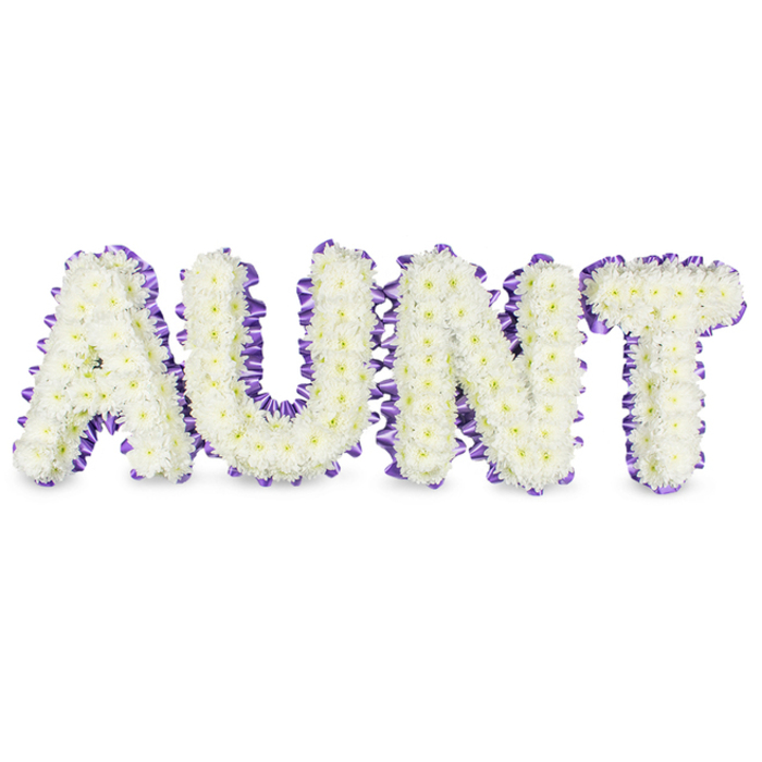 Presented against a clean white surface, the word AUNT is rendered in large, elegant capitals, each letter meticulously packed with small white spray chrysanthemums to form a plush, continuous face of tiny petals. The chrysanthemums' texture is soft and slightly ruffled, their pale yellow centres showing through in places like gentle highlights that prevent the white from feeling flat. Encircling each letter is a luxurious band of deep purple satin ribbon, pleated to create a scalloped or ruched border that gives dimensional contrast and a refined outline to the floral letters. The ribbon's satin finish captures light and provides a visual rhythm along the frame while subtle grey shadows beneath the composition reinforce the solidity and three-dimensional presence of the tribute. The arrangement suggests the intimacy of handmade work: careful gluing or wiring of each stem, precise trimming to achieve uniform contours, and deliberate spacing to allow the chrysanthemums' tiny heads to breathe. There is an understated scent-a fresh, green chrysanthemum note-that lends a living quality to the design without overwhelming. Intended as a memorial or sympathy tribute for funeral services, graveside remembrances, or a family gathering, the white-and-purple palette reads as respectful and consoling, balancing purity with a whisper of colour. Locally, such personalised letters are often chosen by families in the New Barnet area when they wish to honour an aunt with a clearly readable, dignified floral message that carries both tenderness and composure.