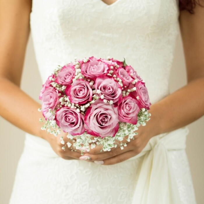 This evocative photo, taken just off the avenues of New Barnet, offers a close-up of a bride (or celebrant) holding a breathtaking, compact wedding bouquet against the soft lace of her sleeveless white dress. The arrangement is a florist's study in contrast and texture: Each rose boasts a gentle blush pink at its centre, blooming outward into saturated fuchsia and magenta, creating a vivid, painterly effect for those admiring each petal's detail. Billowing clusters of baby's breath (gypsophila) weave through the roses, conjuring images of clouds caught in morning sunlight and giving the bouquet its classic romantic softness. Barely visible, fresh green stems hint at careful hand-tying - a signature of bespoke bouquets from Flowers New Barnet. The bride's hands are visible beneath the blooms, their subtle tan and delicate, light-polished nails echoing a sense of understated elegance. The dress she wears is adorned with intricate lacework and a fluttering tie sash, elements that heighten the sense of occasion and tradition. The softly blurred, neutral backdrop suggests a venue steeped in gentle luxury, keeping all attention on the floral artistry at the fore. Altogether, this image speaks to the timeless magic of weddings in New Barnet, offering inspiration for anyone seeking local bridal flowers, meaningful ceremony designs, or simply a celebration of love's beautiful details.