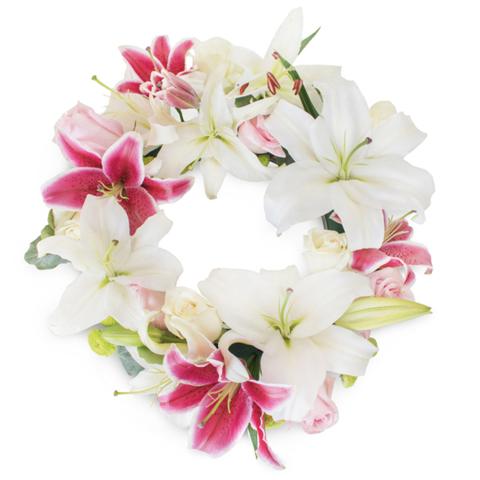 Dominating the foreground is an exquisitely hand-crafted floral wreath, set against a pristine white canvas, evocative of the quiet dignity found in New Barnet gatherings. Cascades of star-shaped white lilies form a luminous core, their petals creamy and pure, each flower opening generously to reveal graceful pistils and gentle green centers-a glowing testament to honour and remembrance. Vibrant fuchsia Stargazer lilies are tastefully woven throughout, their intensity heightened by darker speckles and magenta tones that contrast beautifully with the serene whites. Tucked between the lilies are clusters of pale pink and creamy roses in both bud and bloom, their romantic softness infusing the design with hope and gentle memory. Subtle yet vivid greenery, possibly eucalyptus or seasonal New Barnet foliage, frames and supports each bloom, its cooling tone adding depth and visual texture. The circular shape is full and unbroken, inviting the eye to trace its endless path, symbolic of everlasting love and community support. Locals might recognise this elegant design from services near East Barnet Village or tranquil settings close to Dollis Valley Greenwalk. Perfect for commemorative occasions, this arrangement is lovingly prepared by experienced florists in New Barnet, ensuring every detail expresses both compassion and artistry-a truly dignified tribute for those paying respects in the area.