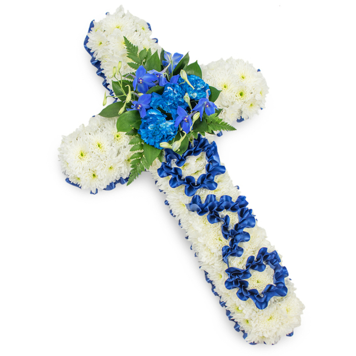 A beautifully composed funeral cross is displayed on a crisp white surface, its outline perfectly proportioned and instantly recognisable as a traditional Christian symbol of faith and remembrance. Every inch of the cross is filled with tightly packed white spray chrysanthemums, their small, rounded petals overlapping to create a smooth, full texture that feels both delicate and reassuringly solid. The glowing whiteness of the flowers symbolises purity and peace, making this a fitting tribute for a father whose memory is cherished. Tracing the entire perimeter, a continuous band of royal blue satin ribbon frames the design, its gentle sheen catching the light and lending a touch of formality and ceremony often seen in tributes delivered to New Barnet churches and chapels. At the centre point, where the arms and stem intersect, a vivid cluster of royal blue blooms forms a dramatic focal feature: ruffled carnations and perhaps blue delphiniums or orchid-like flowers appear in layered tiers, surrounded by rich green fern fronds and glossy leaves that fan outward, adding movement and contrast. Along the vertical length of the cross, just below this central spray, the letters DAD are carefully fashioned from the same royal blue ribbon, looped and folded so that each character stands out clearly against the snowy chrysanthemum backdrop. This simple word, crafted with such precision, gives the piece deep emotional resonance, evoking stories of family life in New Barnet - walks through nearby parks, quiet evenings at home, shared milestones. The overall mood is one of calm dignity and tender farewell, an eloquent floral expression of love, gratitude, and lasting remembrance.
