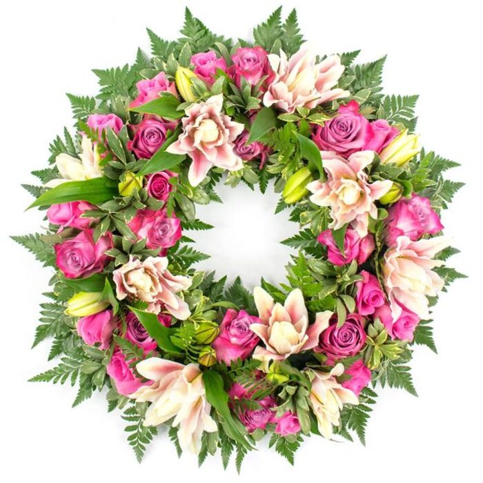 Set against a bright white background that resembles the stillness of a memorial space, this circular floral wreath appears lush and full, its flowers and foliage pressed closely together to form a continuous band of colour and texture. The eye is immediately drawn to the abundance of roses in vivid fuchsia and magenta, their petals richly layered and velvety, creating pools of intense pink throughout the design. Some of the roses are nestled tightly as buds, hinting at life yet to unfold, while others are wide and open, like small, welcoming cups of colour. Interwoven with the roses are large, double-headed lilies whose petals are soft and ruffled, painted in shades of blush, cream, and pale peach, with occasional darker pink tips or veins that give each bloom its own quiet character. A scattering of fresh, lime-green lily buds introduces a note of gentle brightness and suggestion of ongoing life. Surrounding and supporting these flowers is a deep mantle of greenery. Feathery fern fronds extend along the outer edge, offering a delicate, lacy texture, while sturdier, glossy leaves-in varied shapes and subtle tones-fill the inner gaps, ensuring the wreath feels complete from every angle. The overall arrangement is carefully balanced and symmetrical, conveying care and professionalism, much like a bespoke tribute crafted by a local New Barnet florist for a funeral at a nearby church or crematorium. The palette of rich pinks, soft creams, and cool greens, isolated against the plain background, evokes tenderness, remembrance, and a respectful farewell for a loved one.