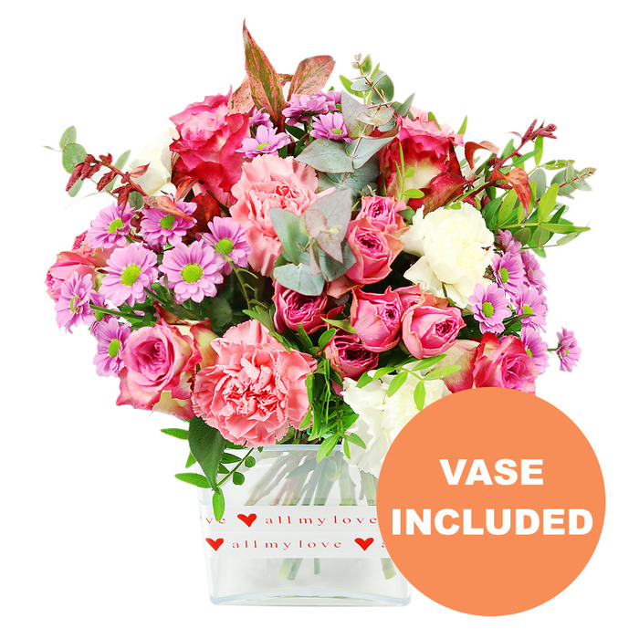A vibrant mixed bouquet, captured against a stark white background, appears as though it has just been carefully positioned on a workbench in a New Barnet flower shop, every stem precisely placed. At the heart of the arrangement are multiple large roses in varying shades of pink, from deep magenta blossoms with gently darkened petal tips to softer, shell-pink blooms unfurling in full, velvety layers. Interspersed with these are generously sized carnations in coral, blush, and peach-pink hues, their ruffled petals forming soft, rounded clusters that give the bouquet a plush, almost cushion-like texture. Weaving between the larger flowers are smaller daisy-style chrysanthemums, some in lilac and lavender tones and others in a more vivid purple, each with a bright green centre that catches the eye and lifts the overall colour palette. Creamy white blooms tucked here and there lend a sense of lightness and help the richer colours sing. Surrounding and supporting the flowers is a lush mix of greenery: rounded eucalyptus leaves with their signature silvery cast, deeper green foliage that frames the flowers, and a few reddish-brown leaves that introduce a subtle, romantic warmth, echoing the mellow tones you might notice in the trees around New Barnet during the cooler months. The entire composition is grounded in a clear, rectangular glass vase with softened corners, giving a clean contemporary look. Around the lower portion of the vase, a crisp white band is wrapped neatly, patterned with repeated red hearts and the phrase all my love, reinforcing the bouquet's affectionate theme. In the lower right corner of the image, an orange circular label clearly reads VASE INCLUDED in white capital letters, highlighting that this is a complete, ready-to-display floral gift, ideal for delivery to homes and offices throughout New Barnet for romantic gestures, family celebrations, or thoughtful thank-you surprises.