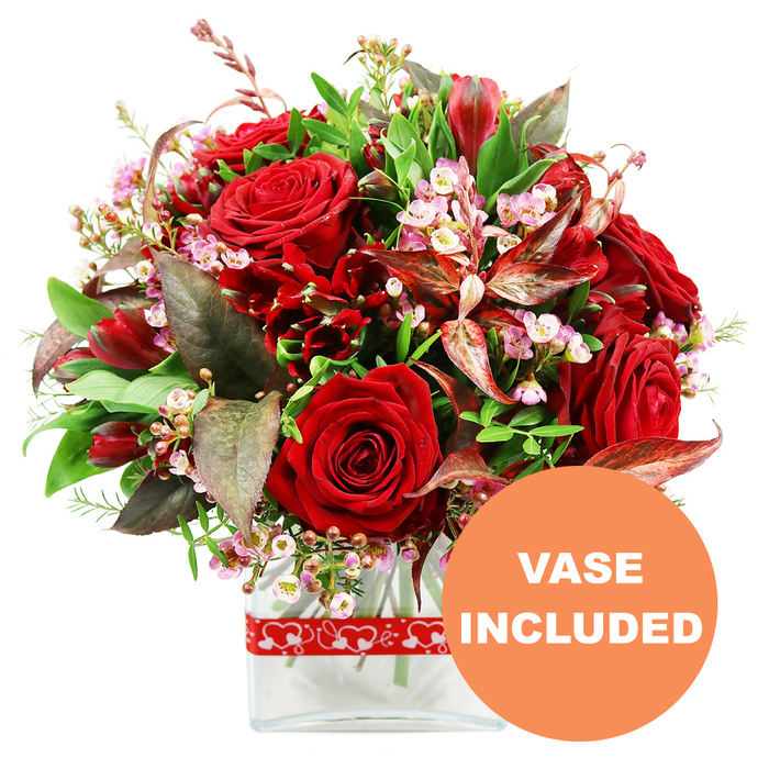 Against a bright white background, a sumptuous red and green arrangement sits in a clear glass vase, as if waiting on the counter of a New Barnet flower shop just off the High Road. At its centre, several large, velvety red roses form the focal point, each bloom at a slightly different stage of opening. Some are tight and bud-like, promising future fragrance, while others have begun to reveal their layered interiors, creating a sense of unfolding romance. Around these roses, clusters of smaller deep red flowers, similar to alstroemeria or compact, multi-petalled blossoms, lend body and intricate texture. Threaded throughout the bouquet are airy sprays of tiny pale pink and white waxflower or limonium, their miniature stars softening the stronger hues and bringing a gentle, almost bridal lightness. Encircling the flowers is a generous collar of foliage: glossy mid-green leaves, finer lighter green sprigs, and distinctive burgundy or reddish-brown leaves with delicate veining that echo the red tones without overpowering them. This mix of foliage adds depth and movement, as if a light breeze were passing through. The bouquet has been shaped into an even, rounded dome, showcasing the practiced craftsmanship of a local New Barnet florist who understands balance and proportion. The stems sit neatly in a modern, clear rectangular glass vase with smooth edges, allowing the green stems and clean water to show through. Around the vase, a red ribbon decorated with neat outlines of white hearts is tied horizontally, reinforcing the romantic message. In the bottom right corner, a circular orange badge with bold white uppercase text announces VASE INCLUDED, making it clear that this is a complete, gift-ready arrangement, ideal for Valentine's Day, anniversaries, or intimate celebrations in New Barnet homes.