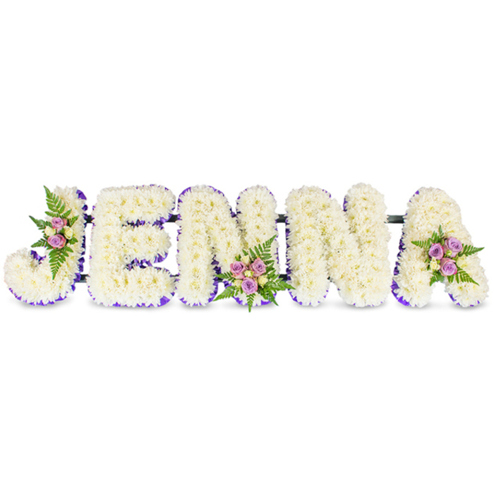 An exquisitely crafted memorial name tribute reads JENNA in five substantial, three-dimensional capital letters, each letter composed of hundreds of small, creamy-white chrysanthemums or similar delicate florets packed densely to create a soft, cloud-like surface. The arrangement sits against a pristine white background so the purity of the blooms and the craftsmanship are plainly visible. A vivid band of deep purple ribbon or a halo of tiny lavender blooms outlines the inner edges and bases of each letter, giving a refined contrast and subtle depth that frames the name with gentle dignity. Three distinct floral clusters accent the piece - one placed on the curving top of the J, a second on the lower centre of the second N, and a third on the upper curve of the final A - each cluster built from soft blush-pink and lavender spray roses, touches of cream or pale yellow roses, and slender, dark-green fern fronds that add a textural, aromatic counterpoint. Thin, dark structural bars link the letters discreetly, providing stability while keeping the display visually cohesive. Soft, directional light brings out the sheen on the ribbon and the layered petals, creating a mood of quiet reverence. As New Barnet's floral specialists, Flowers New Barnet often prepare such personalised tributes for local services - whether at a chapel near Brunswick Park or a committal on a side street off the High Street - offering a refined and heartfelt way to honour a loved one's name with scent, texture, and grace.