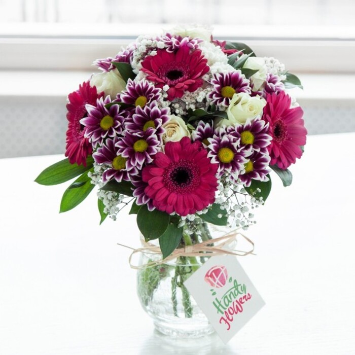 A beautifully composed mixed bouquet is showcased in a low, clear glass vase with a gently textured surface, positioned against a luminous, softly out-of-focus white background that suggests daylight pouring through a large window in a New Barnet living room. Dominant within the arrangement are several striking gerbera daisies in intense shades of deep fuchsia and magenta, their broad, symmetrical petals forming perfect discs around inky, almost black centres that create a strong visual anchor. Surrounding them, bicolour chrysanthemums offer intricate detail: each bloom features layers of narrow petals with rich purple tips that gradually fade into creamy white, gathering around bright yellow centres that add warmth and lift the mood. Interspersed between these larger, showier flowers are clusters of creamy white spray roses, some fully open with soft, spiralling petals, others still in bud, their outer sepals hugging the petals in a promise of continued blossoming over the next few days. Delicate sprays of white baby's breath weave gracefully through the bouquet, lending an airy, cloudlike lightness that softens the bold pinks and purples. Deep green foliage forms a natural framework, the leaves' glossy surfaces catching the light and providing an elegant contrast that highlights the saturation of the blooms. The stems are visible through the vase, neatly arranged in the clear water, hinting at the careful craftsmanship behind the design, much like a bouquet you might order from Flowers New Barnet for same-day delivery around Hadley Wood or Oakleigh Park. Around the neck of the vase, a rustic loop of natural twine is tied in a simple bow, from which hangs a small rectangular gift tag in soft white, decorated with a pink floral emblem and flowing script that suggests a personal message. The entire scene conveys freshness, joy, and the quiet excitement of receiving a thoughtfully chosen floral gift, perfect for celebrating milestones or simply brightening a New Barnet home.