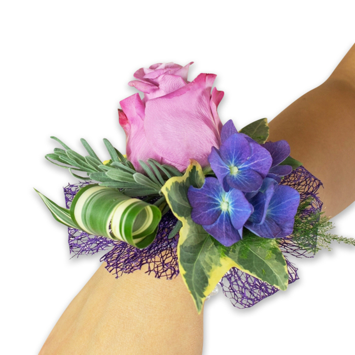 A beautifully balanced wrist corsage is photographed on a light-toned forearm against a stark white studio background, evoking both classic elegance and modern floristry accents often requested by clients from Station Road and nearby Hadley Green in New Barnet. Dominating the arrangement is a large, partially open rose in a soft pink-lavender blend, its outer petals kissed with a deeper fuchsia rim that adds a sense of movement and romantic depth. Flanking the rose are three dense clusters of deep indigo-purple hydrangea, each tiny floret revealing a subtle blue-tinged center that creates a luminous, almost porcelain contrast. Interwoven are long, slender silvery-green sprigs-evoking eucalyptus or lavender-that provide a soft, feathery halo and a faint herbal scent. Glossy variegated ivy leaves contribute classic texture, their dark green cores framed by bright cream edges, while a broad leaf with striking white-and-green striping is worked into an elegant curl, offering a sculptural, contemporary touch. Below the floral elements, a dark purple open-weave mesh peeks through, lending shadow, volume, and a fashionable hint of evening glamour. The colour story-muted mauve, vivid indigo, and silvery foliage-reads as refined celebration; you can imagine this corsage worn at a wedding near New Barnet's local churches or as a finishing detail for prom photographs taken on Barnet High Street. Meticulously assembled, it balances tactile richness and visual drama, ideal for anyone seeking a fresh, handcrafted accessory with a strong local sensibility.