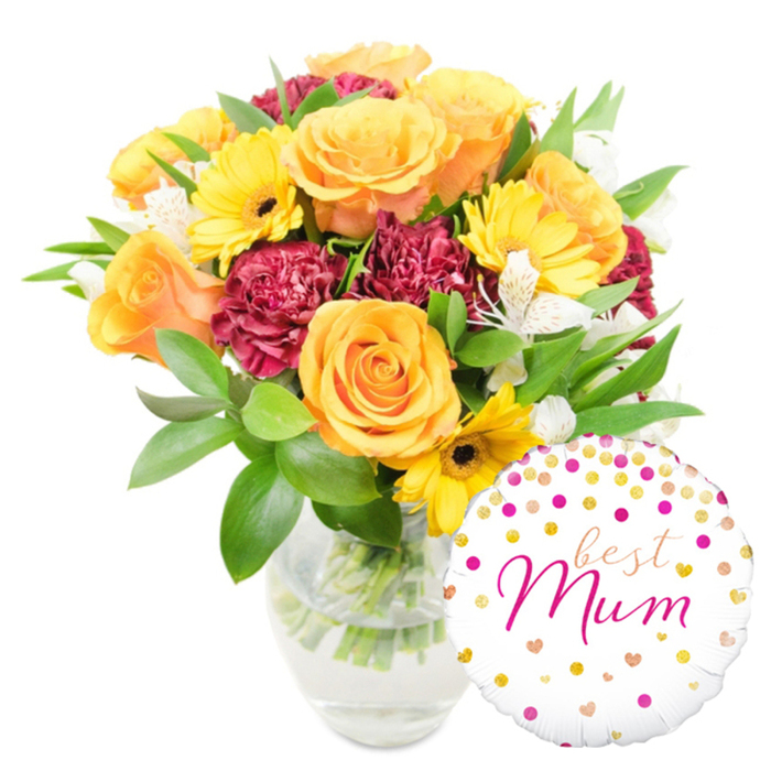 A lively, heartwarming floral gift pictured on a pure white field: clear glass reveals crisp stems and water, anchoring a joyful composition of warm-hued blooms. Prominent are full, orange and yellow roses with gently spiralled petals that catch soft, studio light and give off a mellow floral perfume. Intermingled are bright yellow gerbera daisies whose flat, radiating faces and dark brown centers create cheery focal points, while deep red-to-burgundy carnations lend voluptuous texture with their layered, ruffled petals. Fine sprays of white alstroemeria - delicate Peruvian lilies with slim petals and faint speckling - dance among the bolder flowers, adding lightness and refined detail. The whole is cushioned in abundant greenery: glossy broad leaves that reflect the light and slender sprigs that push between blooms to provide a garden-fresh silhouette. At the lower right, slightly overlapping the vase, a round white foil balloon is playfully placed; it is patterned with scattered gold and fuchsia polka dots and tiny heart shapes and bears the affectionate inscription best Mum in a flowing fuchsia script, making the set unmistakably intended as a loving gift. The composition reads as both elegant and informal - like a bouquet hand-gathered for mum before a morning visit - and its scent profile would be softly sweet with warm rose notes. Locally, this design is a popular choice for customers requesting delivery to homes around Station Road, Oakleigh Park and the New Barnet village area when they want to make a mother feel seen and celebrated.