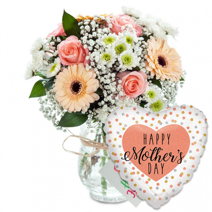 This detailed Mother's Day scene shows a thoughtfully coordinated bouquet and balloon arrangement set against a plain white backdrop, highlighting every colour and curve without distraction. On the left, a lush, rounded bouquet sits securely in a clear glass vase shaped like a classic jar, its smooth sides revealing the stems gathered neatly within and a simple tie of light brown twine looped around the neck. The focal flowers are several fully opened roses in soft peach and blush-pink tones, their ruffled petals gently unfurling to create a romantic, spiral heart at the centre of each bloom. Partnering them are two striking peach gerbera daisies, each with a distinct deep brown centre that draws the eye like a sunlit focal point, reminiscent of bright flower beds near New Barnet's residential streets. Around these larger blooms, clusters of airy white gypsophila create a misty halo, while smaller white chrysanthemums with pale green centres tuck into the gaps, adding shape, fullness, and a subtle hint of spring freshness. Threads of rich green foliage arc between the flowers, some leaves softly serrated, others smooth and rounded, giving a layered, garden-inspired feel. Just behind the bouquet, partly concealed by stems and petals, peeks the corner of a small white greeting card, suggesting a handwritten Mother's Day message from a family not far from Victoria Park. To the lower right, overlapping the vase and drawing the viewer's gaze, a large heart-shaped foil balloon adds a festive flourish. Its clean white surface is dotted with shining gold and silver confetti-like circles, and at its centre a generous peach-coloured heart frames the greeting HAPPY Mother's DAY in bold black type and elegant cursive. The overall effect is warm, joyful, and perfectly suited to Mother's Day flower delivery throughout New Barnet and nearby areas.