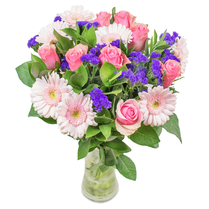 An elegant floral arrangement, crafted in the heart of New Barnet, sits confidently in a slender, clear glass vase against a crisp, white backdrop. The centrepiece features a harmonious mix of 7-9 classic pink roses, many petals still coyly wrapped in tight whorls or just starting to unfurl, displaying their romantic, spiral shapes that capture the timeless beauty found at Victoria Park in spring. Intermingled are 5-7 delicate pale pink gerbera daisies, their broad, cheerful faces and lighter, creamy centres glowing softly in diffused daylight. Clusters of vivid purple statice, their texture distinctive and colour bold, compose gentle arcs throughout the bouquet, highlighting the playful side of local floral artistry. Lush, deep green foliage forms a vibrant, abundant base, enhancing the arrangement's freshness and echoing the leafy oasis of New Barnet's gardens. Water glistens in the vase, accentuating the stems and emphasizing their just-picked vibrance. Ideal for birthdays, anniversaries, or brightening any New Barnet workspace or home, this bouquet beckons with an inviting sense of celebration, affection, and cheer.