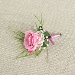 A single blush pink rose boutonniere is displayed on a pale beige, linen-like fabric, its refined simplicity capturing the mood of a romantic wedding morning in New Barnet. The camera looks down from a slightly overhead angle, revealing the rose in a semi-open state, its petals unfurling from a soft, powdery pink center into deeper pink edges that curl back gently, as if caught mid-breath. Each petal appears velvety and fresh, suggesting it has just been conditioned by a careful florist. Encircling the base of the rose are airy tufts of white baby's breath, their countless tiny blossoms creating a misty, cloud-like frame that softens the transition between the rose and the greenery. Fine, grass-like blades of bright green foliage radiate lightly from behind, giving the arrangement a sense of movement and natural grace, reminiscent of leaves rustling along the residential streets near New Barnet station. Nestled beneath, a single, wider dark green leaf is folded with precision, wrapping protectively around the stems and forming a clean, architectural base. The stems themselves are bound with a satin ribbon in shades of blush and light lavender-pink, the sheen catching the light as it is tied into a modest, elegant knot rather than a fussy bow. This subtle ribbon detailing adds a refined accent, harmonising with pastel bridal palettes often chosen for local ceremonies. The softly textured linen ground, with its visible weave and warm neutral tone, allows the pinks, whites, and greens to stand out clearly without harsh contrast. The overall impression is of a thoughtfully designed lapel flower-perfect for a groom, groomsman, or prom date heading to a celebration anywhere across New Barnet and the surrounding North London suburbs.
