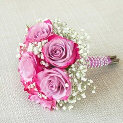 An elegant hand-tied bouquet of roses and baby's breath lies on a pale oatmeal-coloured fabric, its rounded shape carefully composed to feel both luxurious and intimate. At the heart of the design are several full, open roses displaying an exquisite two-tone effect: the outer petals glow in rich fuchsia and magenta, while the inner whorls soften into cool lavender and light purple, creating depth and a painterly play of colour. Each rose looks freshly cut, with velvety petals that almost seem to hold the faint scent of a classic garden rose on a mild New Barnet evening. Nestled between these statement blooms are delicate sprays of white gypsophila, or baby's breath, their tiny, cloud-like flowers adding volume and a feathery counterpoint to the more robust rose heads. The stems are neatly aligned and wrapped in a smooth satin ribbon, possibly in a deep plum or purple shade, which embraces the handle in a tight, clean line. Over the ribbon, several rows of iridescent white pearls spiral downwards, creating a refined, tactile detail that hints at bridal jewellery and heirloom accessories. A small portion of the natural green-brown stem ends is visible beneath the wrapping, grounding the design in its fresh, organic origins. The light beige textile underneath-reminiscent of linen you might find in a stylish New Barnet townhouse or boutique venue-adds subtle texture without competing with the vibrant florals. This bouquet feels made for a wedding aisle, registry celebration, or engagement photos around local landmarks, radiating joy, romance, and the skill of an experienced florist.