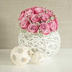 A delicate floral still life captures a rounded bouquet of soft pink roses and whisper-light baby's breath, styled as if for a chic New Barnet coffee table or a welcoming hallway console. The roses are the main focus, gathered closely so that their heads form a smooth, dome-like canopy. Each bloom shows a subtle colour shift, with the inner petals a gentle, almost porcelain blush and the outer edges deepening into a richer pink, approaching fuchsia. Between these roses, clusters of tiny white Gypsophila flowers weave in and out, their airy blossoms giving the arrangement a dreamy, cloud-like halo. The bouquet rises from a distinctive spherical white vase, fashioned from thin, interlaced strands that resemble white-painted rattan or yarn. This openwork lattice creates a sense of lightness, allowing glimpses of shadow and background through its gaps, and lending the design a handcrafted, contemporary feel. Resting on the same white surface, two decorative woven spheres in light, natural wood tones echo the vase's texture in a slightly more rustic way; the larger sphere leans gently toward the smaller, like a carefully considered interior styling detail. Behind everything, a softly textured wall in warm beige or cream works as a quiet backdrop, letting the roses' pink tones and the crisp whites stand out without distraction. The lighting is soft and even, reminiscent of a bright but overcast afternoon in New Barnet, illuminating every petal and weave without harsh contrasts. The arrangement suggests a mood of tenderness and understated celebration, ideal for marking a birthday, new home, or simple thinking of you moment for someone living near High Barnet station or along the leafy streets towards Cockfosters.