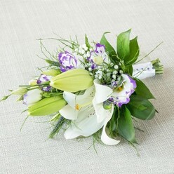 An elongated bridal-style bouquet rests at a gentle angle on a pale beige, linen-like fabric, the neutral background making every flower stand out as clearly as if you were holding it in your hands at a New Barnet consultation. At the centre, several open white Oriental lilies display their full beauty, each petal thick, satin-smooth, and slightly curled at the tips, with golden-yellow stamens rising from the middle and a dusting of warm speckles near the throat. Alongside, you can see firm, lime-green buds that haven't yet opened, giving the arrangement a sense of ongoing life and fresh fragrance. Threaded throughout are lisianthus blooms in white and purple, their ruffled, bell-shaped petals outlined in rich violet and fading softly into creamy centres, bringing a modern twist to the classic lily look. Sprigs of delicate baby's breath weave between the larger flowers, creating little puffs of white that feel like mist on a spring morning walk through nearby Oakhill Park. The greenery is generous yet controlled: shiny, broad dark green leaves provide a solid backdrop, while fine, wispy foliage and a few slender, more upright greens give lightness and movement. The stems are bound together with a smooth white ribbon, neatly wrapped and finished with a line of tiny pearly beads that hint at bridal jewellery and special occasion finery. The overall mood is serene, hopeful, and quietly luxurious, the kind of bouquet you might carry down the aisle in a New Barnet church or registry, or have delivered to a loved one's door to mark an engagement, milestone birthday, or elegant dinner at home.