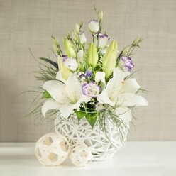 An elegant wedding table arrangement in soft whites and lilacs is captured against a tranquil, linen-textured backdrop, reminiscent of a calm New Barnet afternoon near Victoria Park or the leafy streets off East Barnet Road. At the heart of the image stands a distinctive round white vase, its open-weave, rattan-style lattice lending an airy, sculptural quality, as though the flowers are cradled in a basket of light. From this textured vessel, tall stems of white lilies emerge, some fully open with wide, trumpet-shaped petals and warm pollen visible at their centres, others still sheathed in plump, pale green buds that hint at the bouquet's lasting freshness. Woven among the lilies are graceful white lisianthus blooms, many with petals kissed by soft lavender-purple along their edges or centres, creating a gentle watercolour effect. Some lisianthus buds remain tightly curled, adding a sense of movement and time, as though the bouquet is quietly unfolding. Fine sprays of white baby's breath and tiny, deeper purple accent flowers tuck themselves between the larger blooms, adding depth and a touch of contrast without disturbing the overall calm. Surrounding foliage ranges from feathery, fern-like fronds to broader, rounded leaves in various shades of green, spilling slightly over the edge of the vase to give a natural, garden-fresh feel. On the pristine white tabletop, two woven decorative balls in light beige nestle beside the arrangement, echoing the vase's texture and adding a modern, minimalist touch. Soft, even lighting bathes the scene, allowing the whites to glow and the lilac tones to stay delicate and soothing. The result is a refined floral display that would feel perfectly at home on a New Barnet wedding table, at an intimate celebration in a local restaurant, or as a thoughtful delivery to someone in need of quiet comfort.