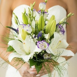 A radiant morning in New Barnet finds a bride holding her floral masterpiece-an exquisite bouquet designed by the hands of skilled local florists who understand the nuances of wedding day emotion. The bouquet captures the eye immediately with large, gracefully opened Oriental lilies in pure white, their petals soft and slightly dappled, reminiscent of the gentle sunlight filtering through Hadley Common in early spring. Nestled beside them are elegantly ruffled white lisianthus, delicately edged in lavender purple, adding a hint of whimsy and fairytale allure. Some of these blossoms are still in tight green buds, promising days of beauty yet to unfold. Deep purple, spiky Veronica flowers weave through the bouquet, introducing richness and movement, while delicate sprays of baby's breath seem to suspend each bloom in place, lending a dreamy, ethereal quality. The foliage cascades outward-broad green lily leaves, tender unopened buds, and whisper-thin asparagus fern-gracefully framing the arrangement and lending an organic, garden-picked freshness so familiar in New Barnet's green spaces. The bride's neatly manicured hands, brushed in a subtle neutral polish, cradle the bouquet as if holding both memory and hope. Carefully composed for a wedding ceremony in a local church or garden, this bouquet reflects the understated luxury and natural beauty esteemed across New Barnet, making it an unforgettable choice for brides seeking both elegance and heartfelt romance.