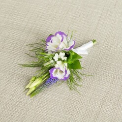 A refined floral boutonniere lies carefully arranged on a pale beige, slightly nubbled fabric that looks like fine linen, its subtle weave adding a soft, tactile backdrop without stealing attention from the flowers. Two white lisianthus blooms sit at the centre, their petals gently curling and folding in on themselves, each fluted edge brushed with a vivid band of purple that deepens into inky tones at the very rim. They look freshly opened, their centres just visible, as if cut from a spring garden bordering one of New Barnet's quiet residential lanes. Nestled among them are airy clusters of delicate white filler flowers, similar to baby's breath, their tiny stars creating a light veil that softens the transition between blooms and greenery. Rising alongside is a slender spike of muscari, its tiny, deep purple, bell-like florets stacked tightly from base to tip, adding both height and a cool-toned contrast. Two pale green buds, still tightly closed, peek from the side, bringing a sense of movement and time-today's celebration, tomorrow's blossoms. Surrounding everything, fine, feathery fronds of greenery spread out like soft lace, adding texture and a slightly wild, garden-picked character, complemented by a few slightly broader leaves that anchor the design. At the base, the stems are gathered cleanly and wrapped in a narrow band of smooth white satin ribbon, neatly finished to create a tidy handle ready for pinning to a jacket. The overall mood is calm, graceful, and quietly romantic, exactly the sort of elegant buttonhole a groom or prom guest might collect from a trusted local florist in New Barnet before heading to a nearby venue or family gathering.