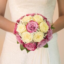 At the heart of New Barnet, this bouquet, held delicately at waist level by a bride in an embroidered ivory gown, reveals a composition that celebrates both tradition and the vibrant local spirit. The bouquet forms a tight circle of creamy lemon-yellow roses, each one thoughtfully chosen for its freshness and gentle hue-a nod to morning walks through Victoria Recreation Ground. Surrounding these are glossy fuchsia-pink roses, some with petals tinged in soft lavender or dusky mauve, echoing the natural transitions seen in local cottage gardens. Sprigs of baby's breath, airy and pure, thread between the blooms, creating a dance of texture and lightness, while deep green leaves peek from the shadows, anchoring the arrangement with a rich, earthy contrast. The bride's bare forearms, visible and steady, cradle the bouquet as if holding a cherished memory; the fine lace overlay and smooth satin panels of her dress shimmer in bright natural light, their intricate details reflecting care and artistry. The softly blurred, pale background highlights the hand-tied florals, inviting the viewer into a moment suspended between joy and anticipation. This arrangement, designed for a romantic celebration in New Barnet, evokes the local charm Flowers New Barnet is known for-an homage to the nuanced art of floral storytelling, perfect for couples or anyone seeking wedding inspiration that captures the area's inviting warmth and sophistication.