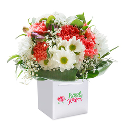 A captivating bouquet in a crisp white gift bag stands as a beacon of celebration and affection, meticulously assembled by skilled florists in New Barnet for heartfelt gifting. Shades of coral and vibrant red carnations, their ruffled blooms lush and inviting, mingle seamlessly with cheerful white daisies-each with a vivid green-yellow center reminiscent of the energetic community spirit found near the high street. Wisps of baby's breath float effortlessly amid the arrangement, their fine, cloud-like blossoms adding a touch of innocence and breathy lightness. Surrounding the florals is a cascade of deep green leaves, broad and glossy, fanning out with a sense of protective grace and reminiscent of the area's tree-lined avenues. Hints of purple-tinted buds peek invitingly from the ensemble's left side, suggesting new beginnings and adding a subtle intrigue only a trained eye might appreciate. The white gift bag, with its strong ribbon handles, sits as a pristine base, perfect for swift, secure delivery anywhere in New Barnet, from Lyonsdown to Oakleigh Park. Light spills softly over the scene, amplifying the bouquet's freshness and the message of joy that comes with each thoughtfully placed stem. This arrangement, so full and radiant, is designed not just to catch the eye but to lift spirits-marked by its ready-to-gift presentation and the unmistakable care of a local New Barnet florist. Ideal for commemorating life's special occasions or offering comfort and cheer in times when heartfelt connection matters most.