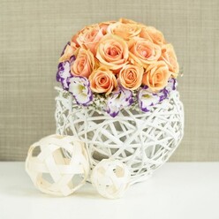 On a smooth, white tabletop, a meticulously arranged floral centerpiece captures the gentle romance of a New Barnet celebration. The composition is dominated by a lush dome of peach and apricot roses, each one fully open and perfectly cupped, their petals layered tightly so the blooms appear almost seamless. The colours shift subtly from soft blush at the outer edges to deeper apricot toward the heart of each rose, creating a warm, inviting glow that feels ideal for weddings or anniversaries. Nestled around the base of this rounded cluster is a ring of white lisianthus, their petals finely ruffled and carefully edged in a deep violet or purple, adding a sophisticated, painterly contrast. Threads of delicate white gypsophila peek between these blooms like tiny constellations, lending airiness and a sense of movement. The entire bouquet is anchored in a broad, spherical basket-style vase made from interwoven white rattan or wicker. Its open latticework shows glimpses of shadow and light between the strands, introducing texture that feels both modern and naturally handcrafted. In the foreground, two pale wooden decorative orbs, one medium and one smaller, rest casually on the same white surface, echoing the woven pattern of the vase and reinforcing the natural, organic theme. Behind, a softly textured beige fabric backdrop, similar to fine linen or burlap, provides a warm, unobtrusive setting that lets the flowers take centre stage. This image suggests a serene, thoughtfully styled table at a New Barnet home close to Station Road or a boutique reception venue near Oak Hill Park, designed to offer guests a sense of calm celebration and quiet luxury.
