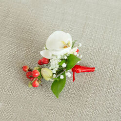 An artfully composed floral boutonniere lies centered on a pale beige, woven linen-style background, its subtle texture echoing the relaxed but refined character of weddings and celebrations around New Barnet. The piece is captured from above, so every element is crisply visible: a flawless white Phalaenopsis orchid forms the central focus, its broad, velvety petals unfurling in gentle curves. At the orchid's core, the throat glows softly with creamy yellow and touches of warm orange, adding depth and a hint of exotic luxury. Encircling this main bloom, clusters of dainty white baby's breath gather like floating clouds, their tiny, star-shaped flowers softening the composition and catching the light in delicate highlights and shadows. Along one side of the design, a tight grouping of shining, cherry-red berries brings a joyful pop of colour, while a few lighter green berry buds interrupt the red with a note of freshness and early-spring energy. Around and beneath the flowers and berries, glossy green leaves in various shapes-some broader and pointed, others slender and elongated-fan out discreetly, framing the orchid and providing a lush foundation. The stems are neatly secured with a bright red satin ribbon, wrapped smooth and finished in a small knot with short trailing ends that echo the berries' richness. This careful ribbon work suggests the experienced touch of a professional florist preparing the piece to be pinned to a jacket at a New Barnet registry office ceremony, a formal dinner near Barnet High Street, or a festive winter ball. The overall effect is one of poised elegance and intimate celebration, a small floral accessory that feels deeply personal and thoughtfully crafted.