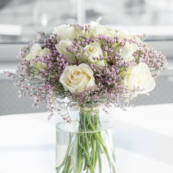 This image shows a graceful vase arrangement of white roses and delicate purple filler flowers, photographed in gentle, natural light that suggests a calm, indoor space somewhere in New Barnet, perhaps a quiet front room off the High Street. At the heart of the bouquet, dozens of roses open fully, their petals layered in classic spirals that create a sense of depth and softness. The colour is a refined creamy white with the faintest suggestion of ivory at the centres, evoking purity, respect, and quiet support. Between these larger heads, airy stems of tiny lavender and blush-pink blossoms weave in and out, forming a halo of pastel colour around the roses. These filler flowers, papery and fine, introduce a gauzy texture that softens any hard line in the design, giving the arrangement a cloud-like, almost dreamlike quality. The clear cylindrical vase is simple and unadorned, allowing the eye to follow the clean, green lines of the stems as they descend into clear, fresh water. Light glances off the glass and the reflective white tabletop beneath, adding small highlights that make the entire bouquet feel newly arranged, as though it has just been delivered to a family home near Victoria Park or a peaceful service in the New Barnet area. The softly blurred background, all pale neutrals and diffused brightness, ensures that the flowers themselves are the only focus. The mood is hushed and tender, a bouquet clearly intended to bring solace and gentle condolence during times of loss.