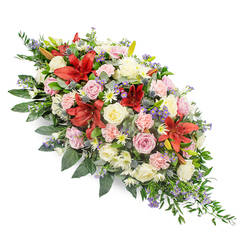 A dignified casket spray stretches gracefully across a white backdrop, its elongated shape echoing the quiet formality of a funeral or memorial service. The eye is first drawn to the rich red lilies at the heart of the composition, their satin petals arching outward like open hands, offering strength and support at a time of loss. Nestled around them are layers of cream, blush, and soft pink roses, some still in bud, others fully unfurled, each one suggesting a cherished memory or a gentle farewell. Light pink carnations, with their frilled edges, mingle with small white daisy-like or chrysanthemum blooms, creating a soft, cloud-like fullness that feels protective and warm. Cool touches of blue-purple thistle and delicate lavender filler flowers weave discreetly through the warmer tones, adding depth and a quiet hint of the unexpected, much like the mix of old and new you notice strolling between New Barnet Station and the local residential streets. A lush base of deep green foliage - glossy leaves paired with featherier, fern-like greenery - frames the spray and extends slightly at each end, making the arrangement appear both abundant and carefully composed. The colour palette of reds, pinks, whites, and purples feels balanced and calming, ideal for resting atop a coffin or displayed near the front of a chapel in New Barnet. This thoughtfully crafted design conveys compassion, remembrance, and a sense of peace, as if created by a local florist who understands the quiet comfort flowers can bring to families during their hardest goodbyes.