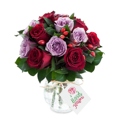 A lovingly arranged round bouquet where deep crimson and burgundy roses mingle with pale lavender and mauve varieties, creating a gentle dialogue of warm and cool tones. Each rose is open and lush, petals layered and slightly ruffled at the edges, suggesting recent harvest and careful handling. Interspersed among the roses are clusters of dark green foliage-likely Ruscus or Salal-whose slender, glossy leaves push back the colour and introduce visual breathing space. Bright red-orange berries, probably rose hips, are dotted through the composition like tiny lanterns, adding a rustic texture and a lively burst that keeps the eye moving across the arrangement. The flowers sit in a reflective mercury-effect glass vase with a rounded, bulbous body and a slightly narrower neck; its antiqued silver surface shimmers in the light and lends an heirloom quality. Around the neck a simple piece of natural raffia or twine is knotted, the finish casually elegant, and a white rectangular tag hangs forward, intentionally minimal so the bouquet reads as an intimate, personal gift. Photographed on a pure white background, the colours sing with clarity and freshness, and one can almost sense the mingled aromas-the deep, velvety warmth of red roses balanced by the faint, sweet powder of lavender blooms and the green, resinous note of foliage. This arrangement evokes romance, thoughtful gifting, and quiet celebration, a floral composition suitable for deliveries across New Barnet and nearby Oak Hill Park when you want to mark an anniversary, birthday, or to simply say you care.