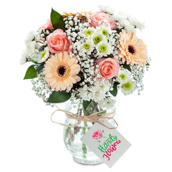 Picture the soft morning light streaming through the windows of a New Barnet home as you receive this artfully arranged bouquet-a gift that captures both elegance and heartfelt emotion. Centered in the design, plush, peachy-pink roses unfurl with classic grace, their velvety petals radiating warmth and tenderness, perfect for expressing affection or comforting solace. Bold, peach-toned gerberas punctuate the bouquet with playful optimism and gentle strength, their wide petals echoing the brightness of bustling High Street mornings. Lush clouds of baby's breath wind between the larger blooms, their tiny blossoms adding a cloud-like softness and whimsical texture to the scene, while clusters of white chrysanthemums, highlighted by fresh green centres, evoke the leafy serenity of Victoria Recreation Ground. Stems of deep green foliage-possibly eucalyptus-peek through, adding structure and grounding aromas reminiscent of a walk along Barnet Road. Every element is presented in a clear, rounded vase, the stems visible and fresh-an assurance of quality sourced daily by New Barnet's experts. Securing the neck, a twine tie finishes the ensemble, adorned with an elegant, empty tag that invites a message unique to the occasion. Created for those special celebrations or everyday surprises, this bouquet is ready for timely delivery across New Barnet, handcrafted with pride for the people who call this community home.